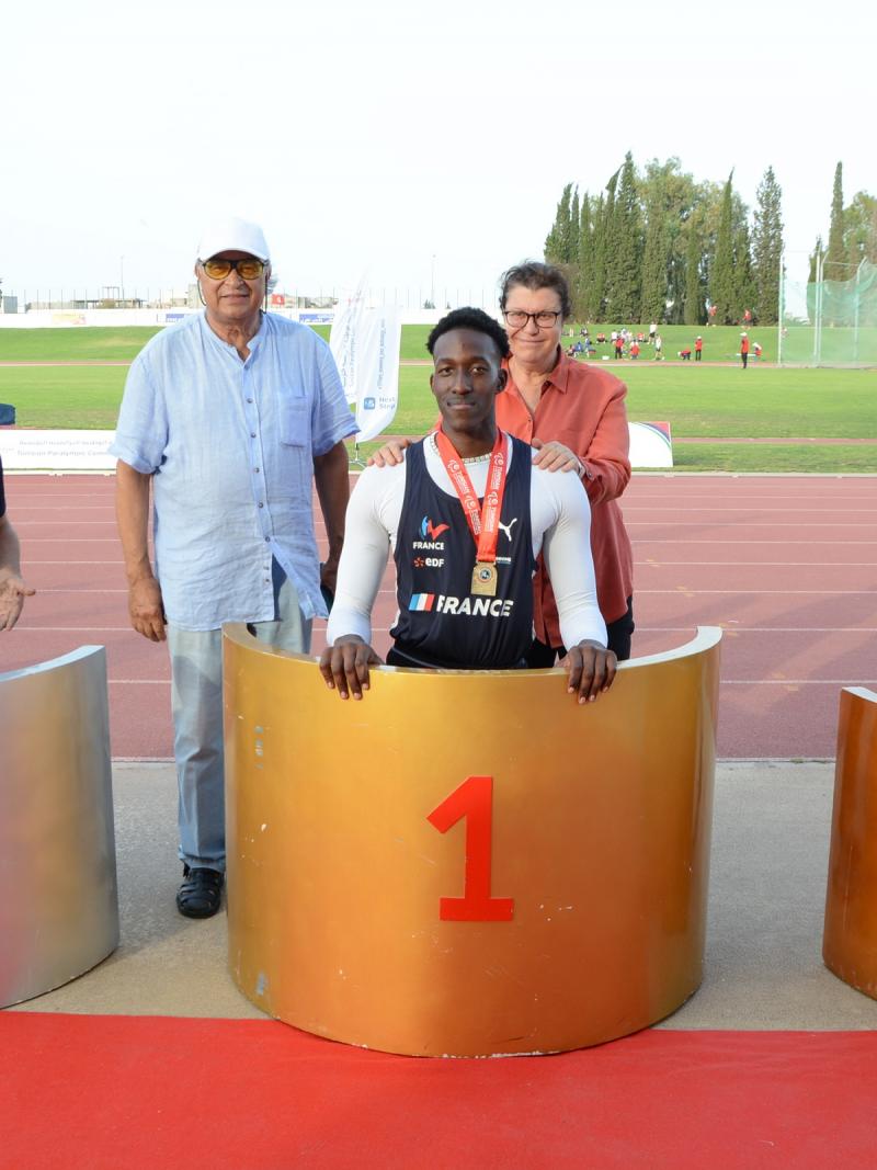 Three men on a podium with a man and a woman in the background on an athletics track