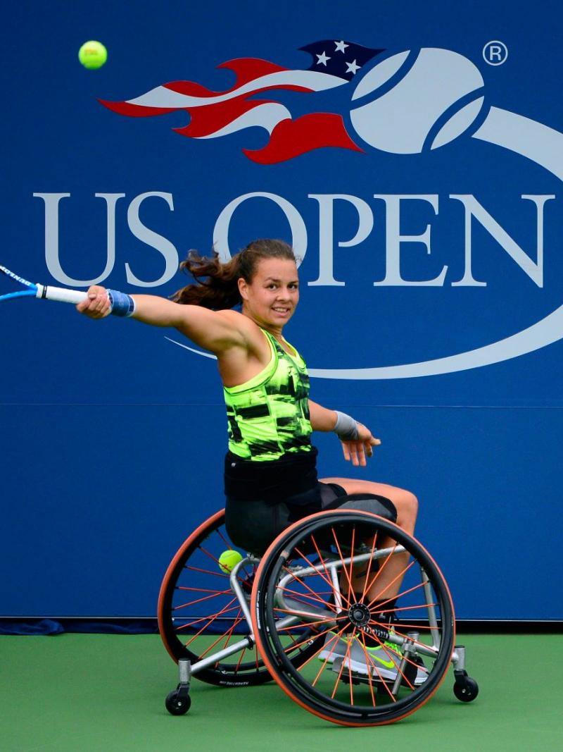 A female wheelchair player returns a shot with the words US Open written prominently on an official backdrop behind her.