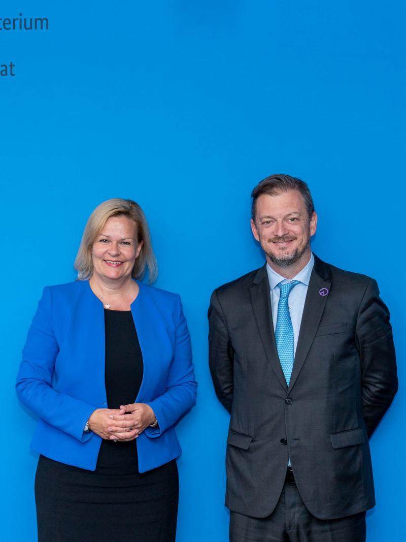 A woman and a man in business attire stand in front of an official blue backdrop.