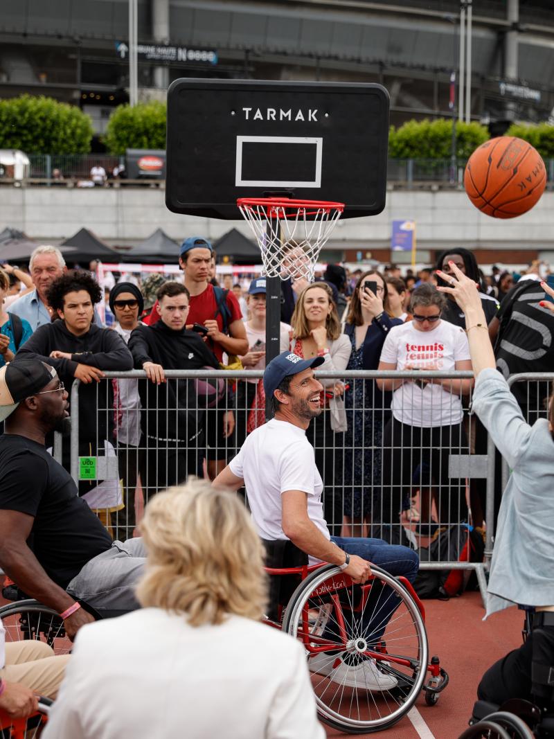 A woman sits in a wheelchair as she makes a basketball shots while a crowd of spectators looks on.