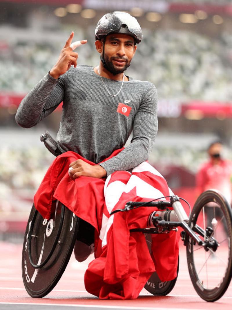 A wheelchair racer, his race chair draped in a Tunisian flag, lifts his hand in celebration on the track.