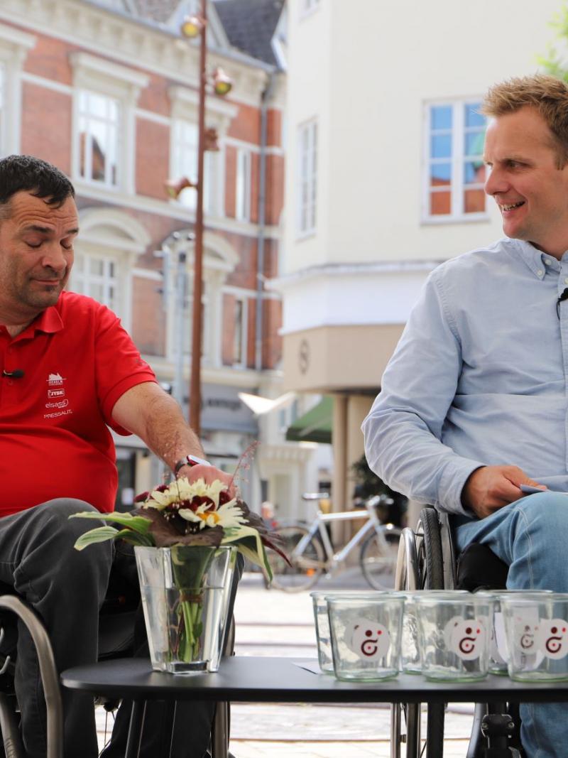 Two men in wheelchairs face a table set with glasses for the 2022 Wheelchair Rugby World Championship draw.