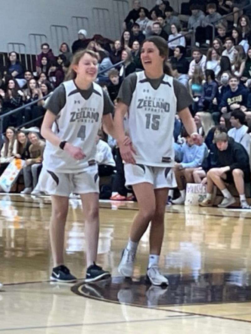 A friend guides a blind girl, both in basketball uniforms, across a basketball court in a packed stadium.