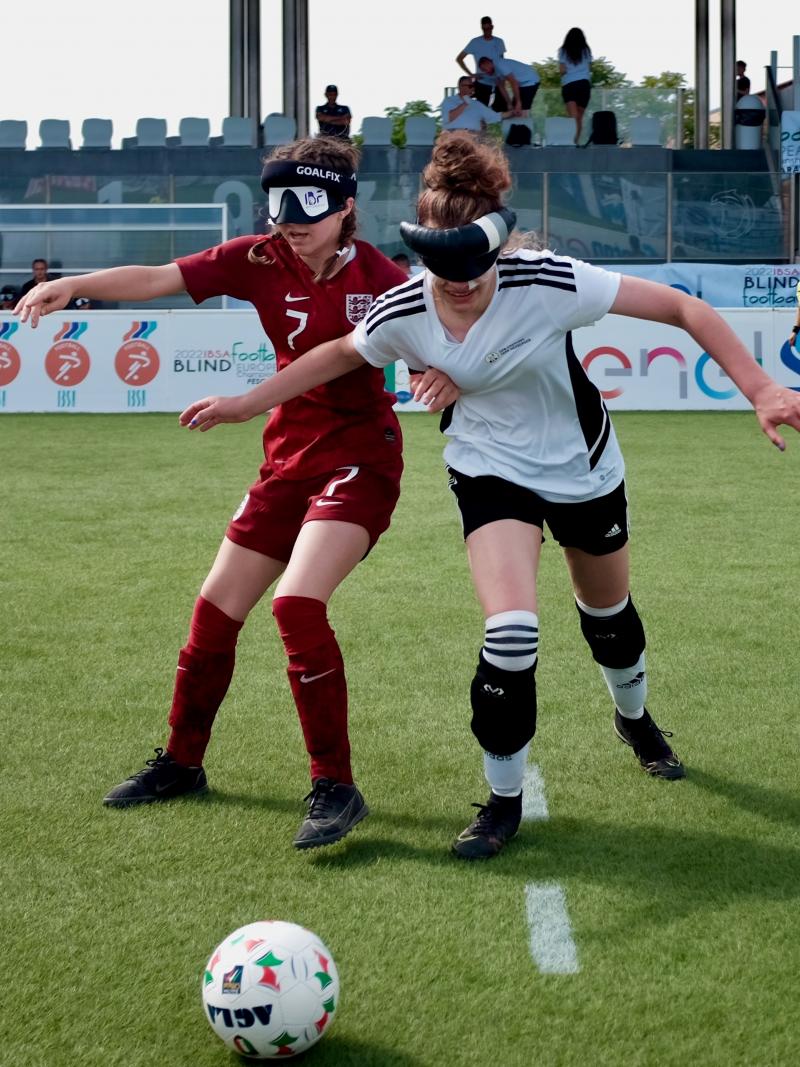 Two female players from different teams chase a ball across a football pitch.