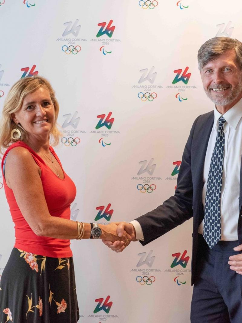 A woman and a man shake hands in front of a Milan Cortina 2026 media backdrop