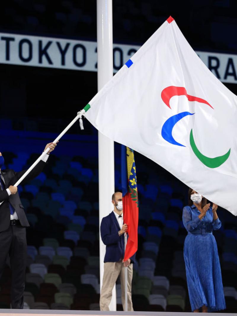 A man waves a flag with the Agitos logo on a stage at a stadium as a man and woman look on.