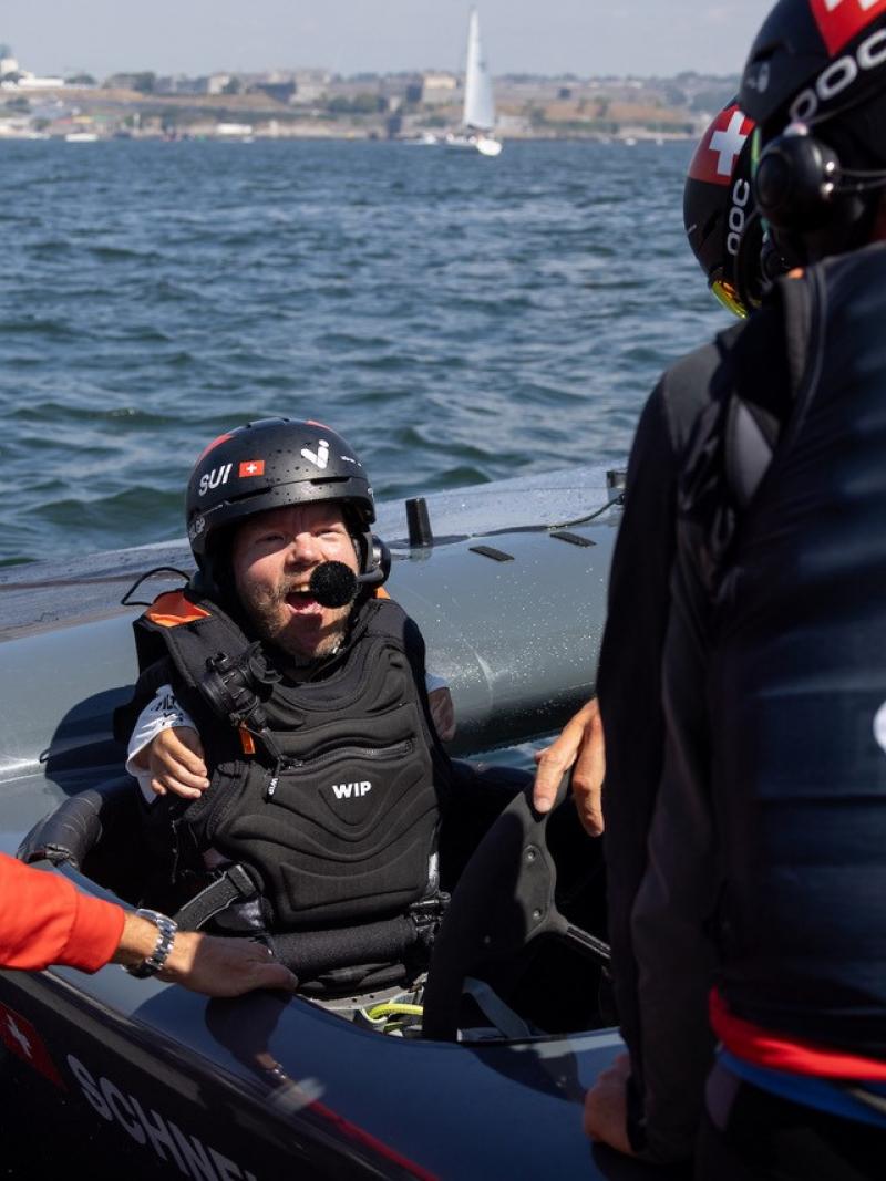 A man in a lifejacket smiles widely inside a sailboat.