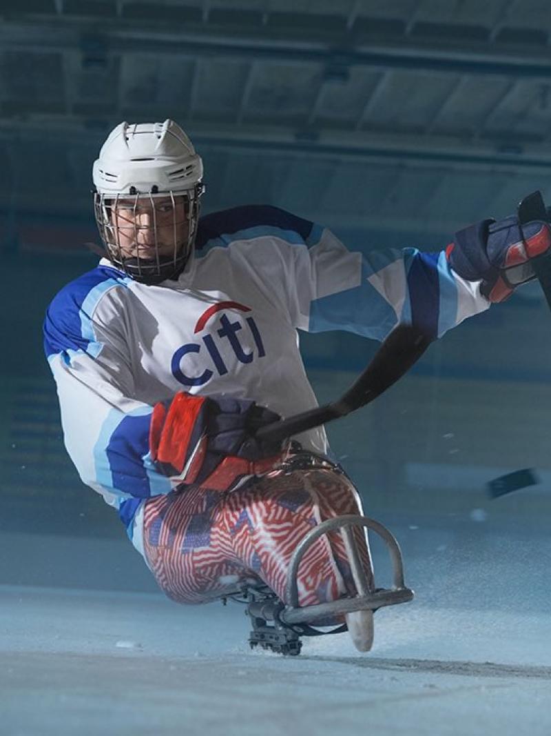 A female Para ice hockey player taking a shot alone in an ice rink
