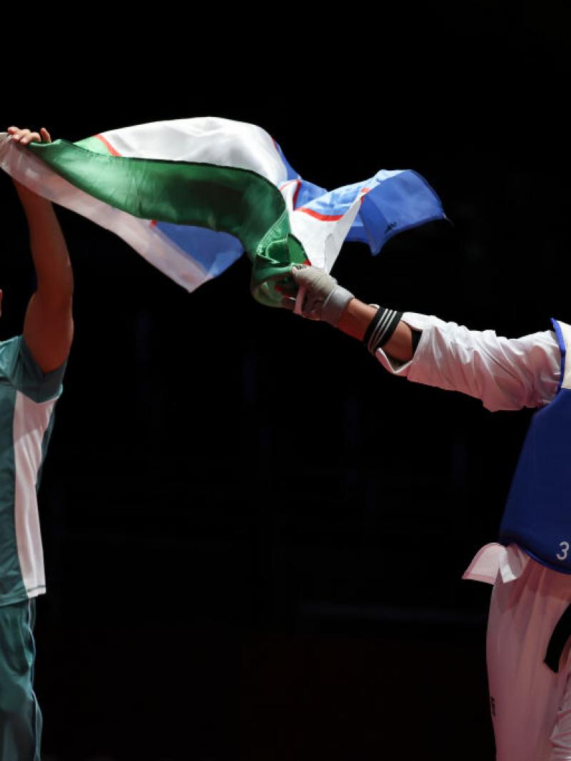 A smiling male coach and female athlete wave an Uzbek flag between them after a competition bout.