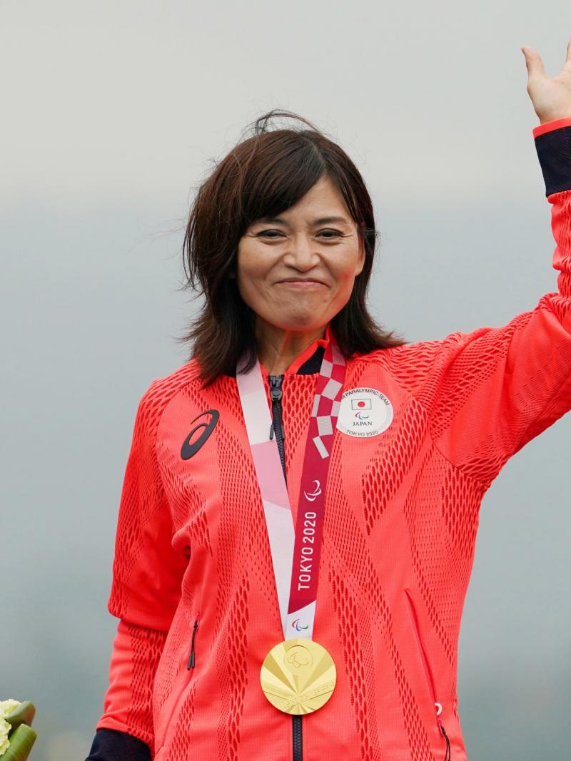 A Japanese female athlete with a gold medal around her neck waves her hand.