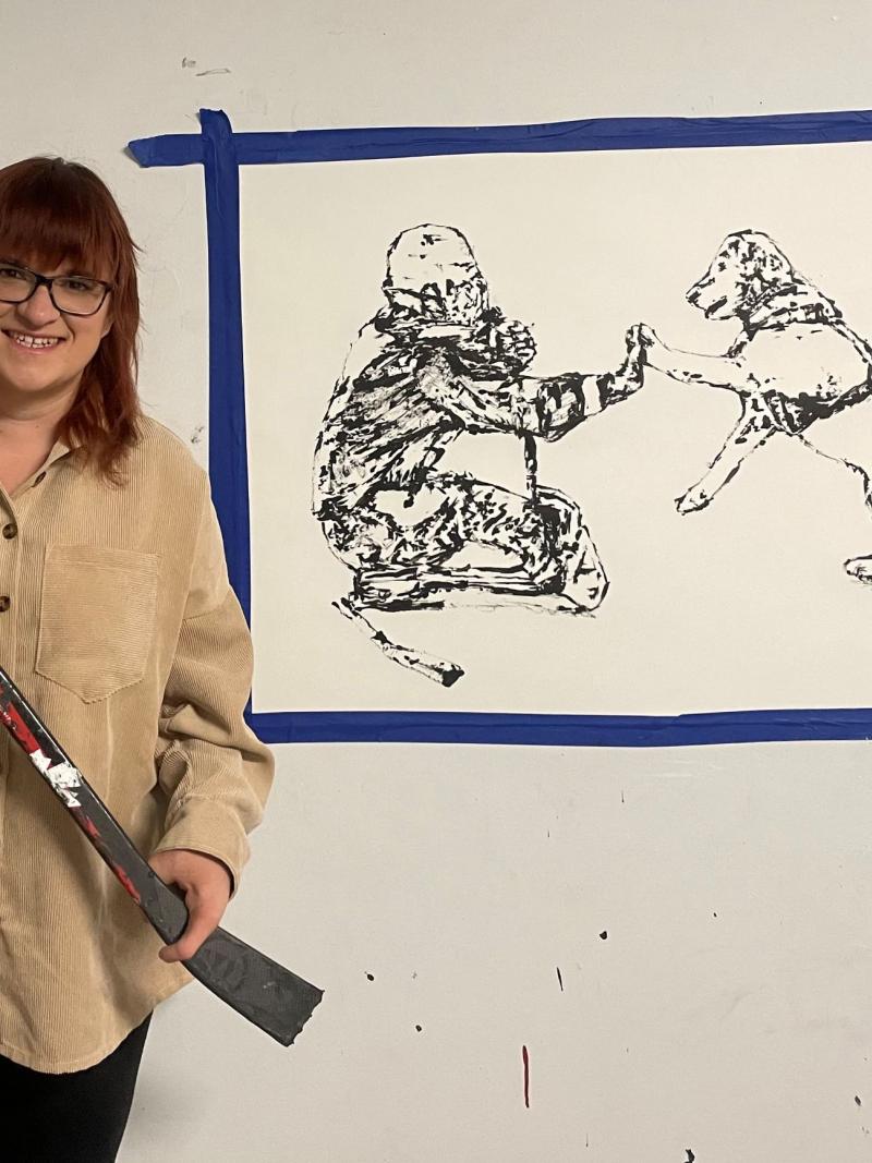 A female artist holding a Para ice hockey stick and stands in front of a painting of a female Para ice hockey player and her dog.
