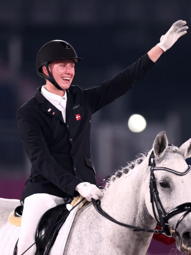 A male athlete riding a horse raises his left hand. He is accompanied by a woman wearing a red and white face mask.