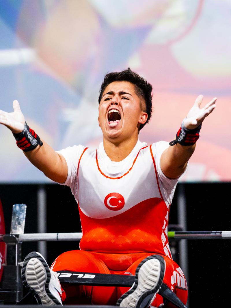 A female powerlifter raises her hands and yells in celebration while seated on a bench.