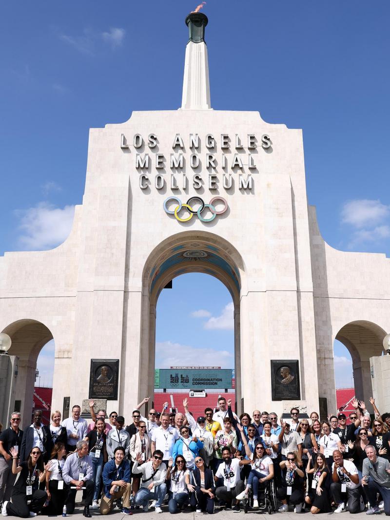 A group of people, many wearing LA28 shirts, pose for a photo in front of the Los Angeles Memorial Coliseum.