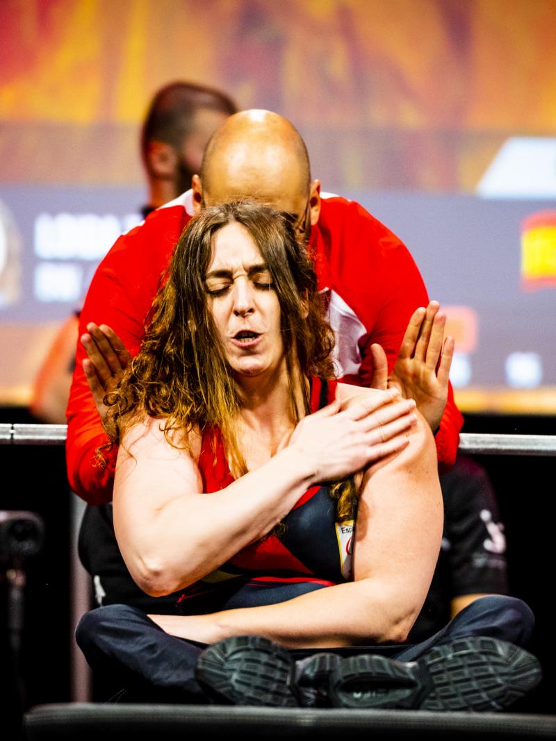 A female powerlifter on the bench as her coach gives her a last minute shoulder warm-up.