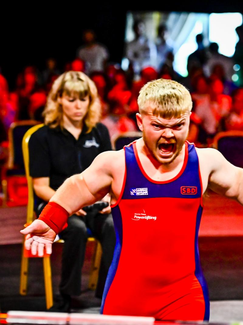 A male Para powerlifter yells in front of the barbell with his arms wide.