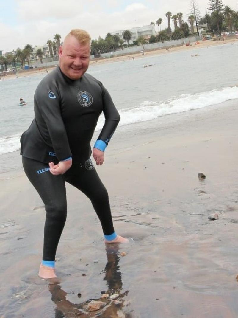A male in a wet suit stands on the ocean shore, his feet in the water.