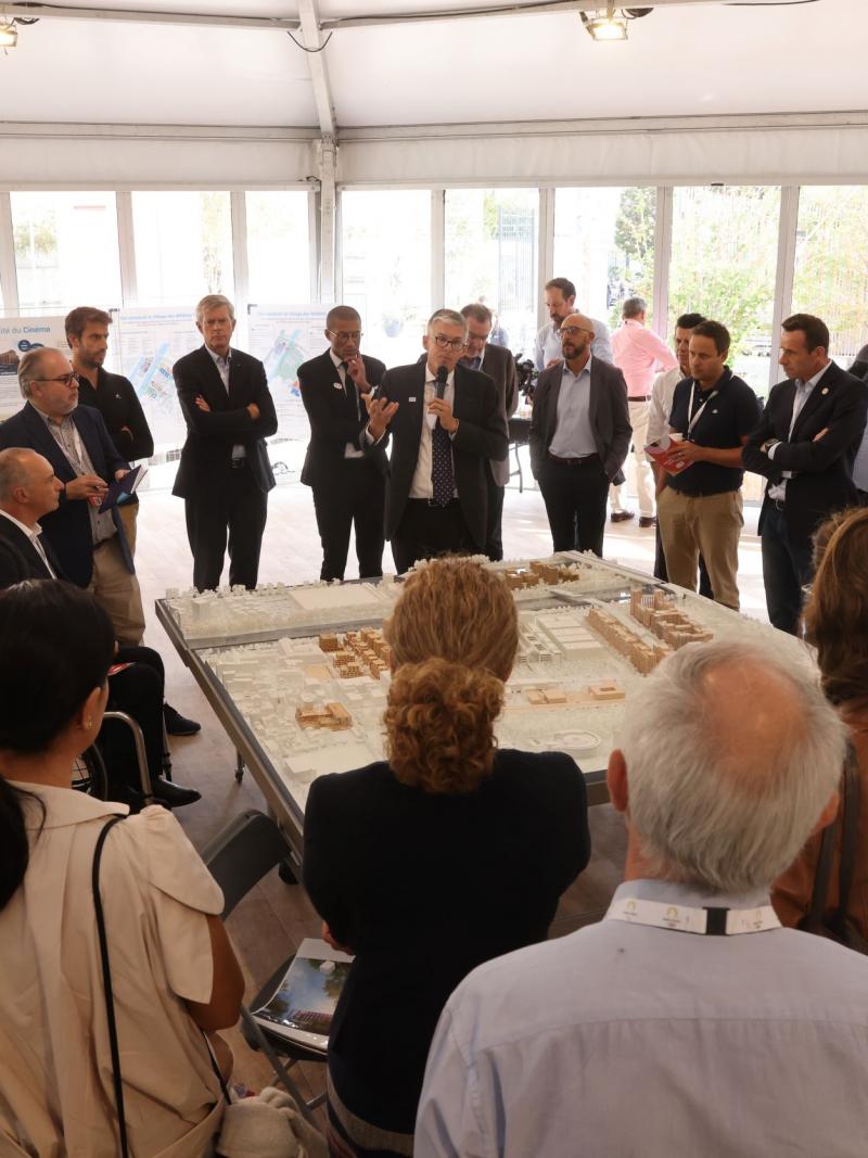 Officials look at a miniature model of a city landscape while one man speaks in a microphone.