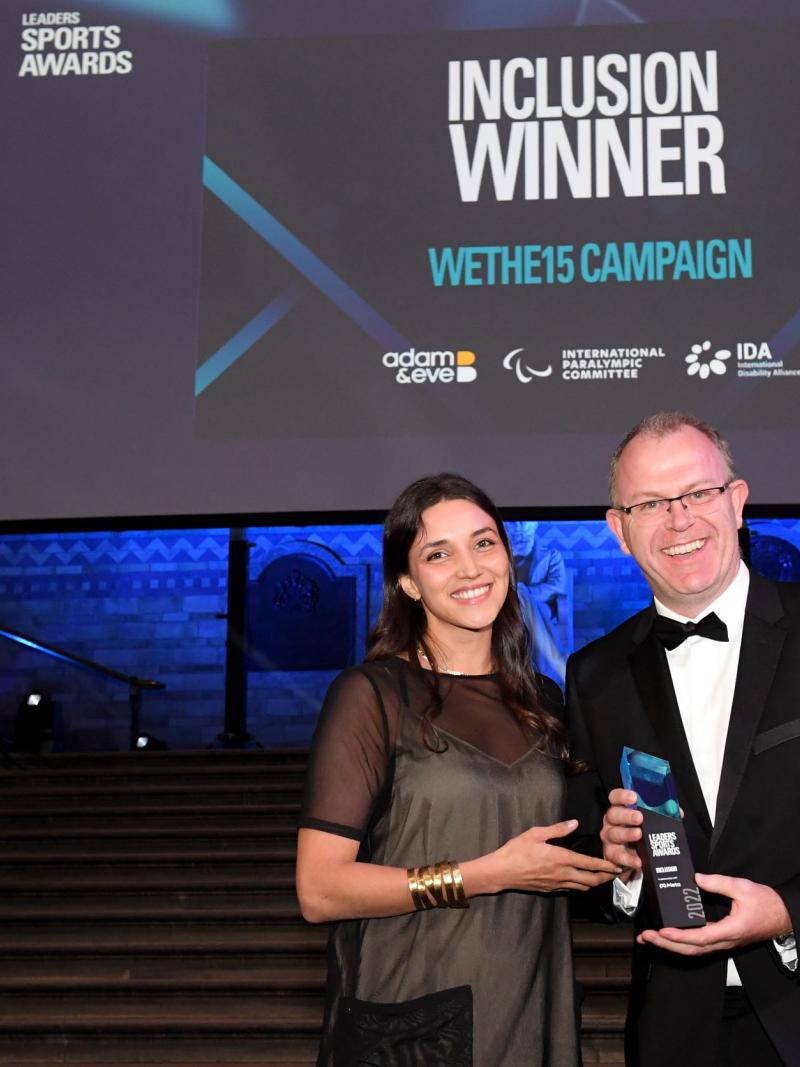 A man and a woman hold a trophy in front of a screen with the words "Inclusion Winner WeThe15 Campaign"