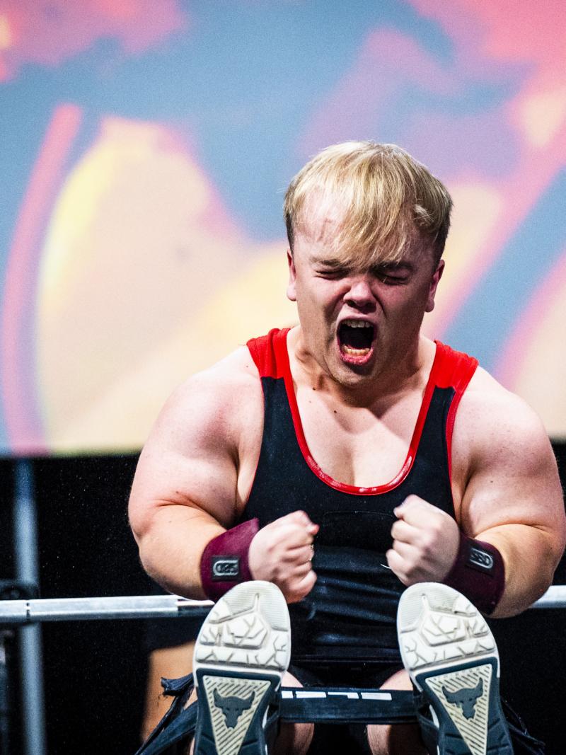 A male Para powerlifter of short stature screams and squeezes his fists in celebration after making a successful lift.