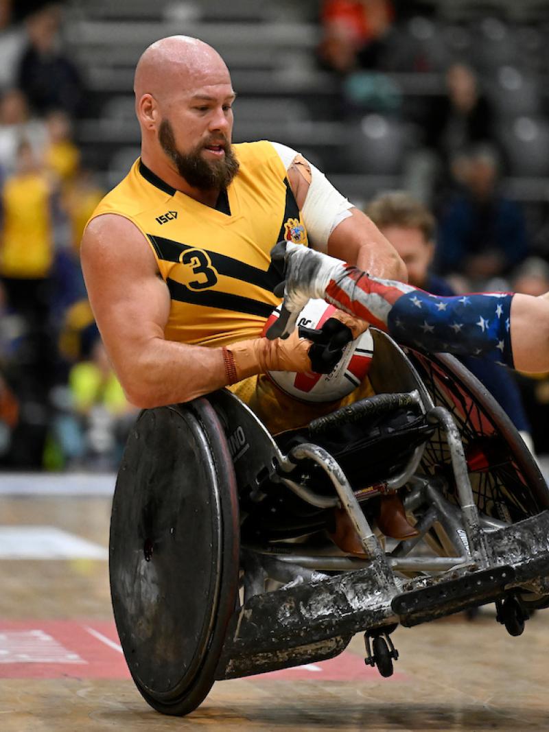 A male wheelchair rugby player wearing a yellow jersey holds onto a ball, while another athlete wearing a blue jersey tries to grab it