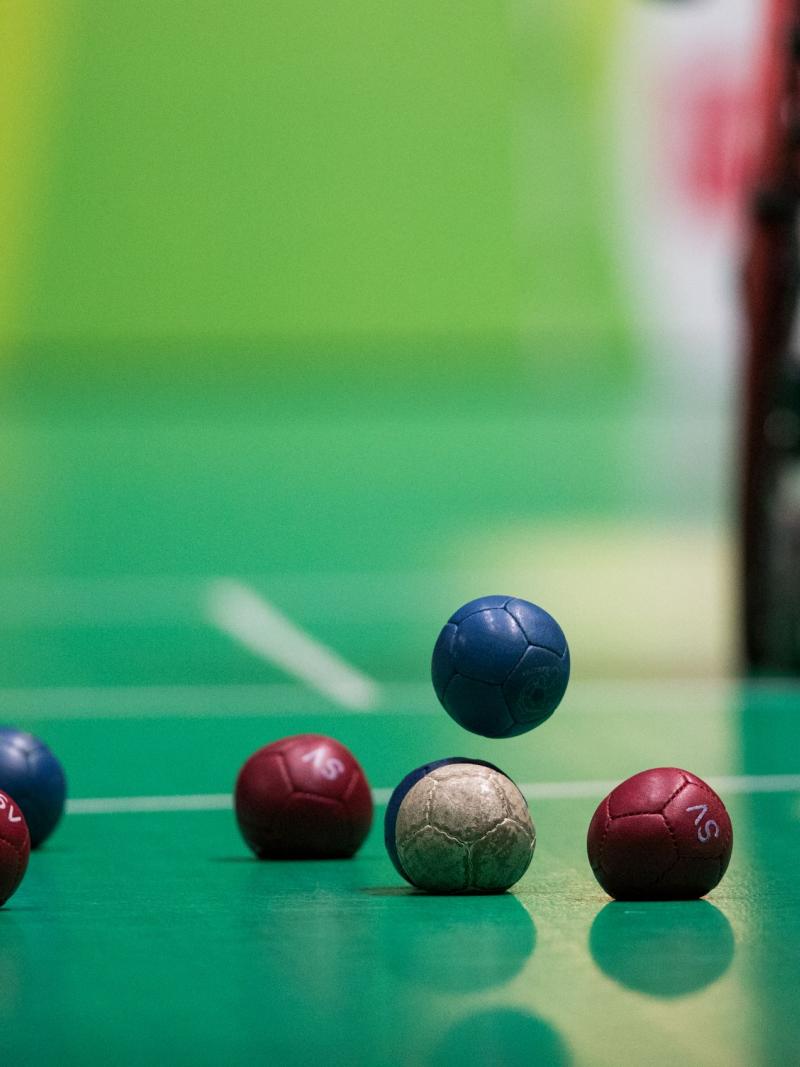 Seven boccia balls lying on the playing ground while one blue ball is falling down, about to touch the ground.