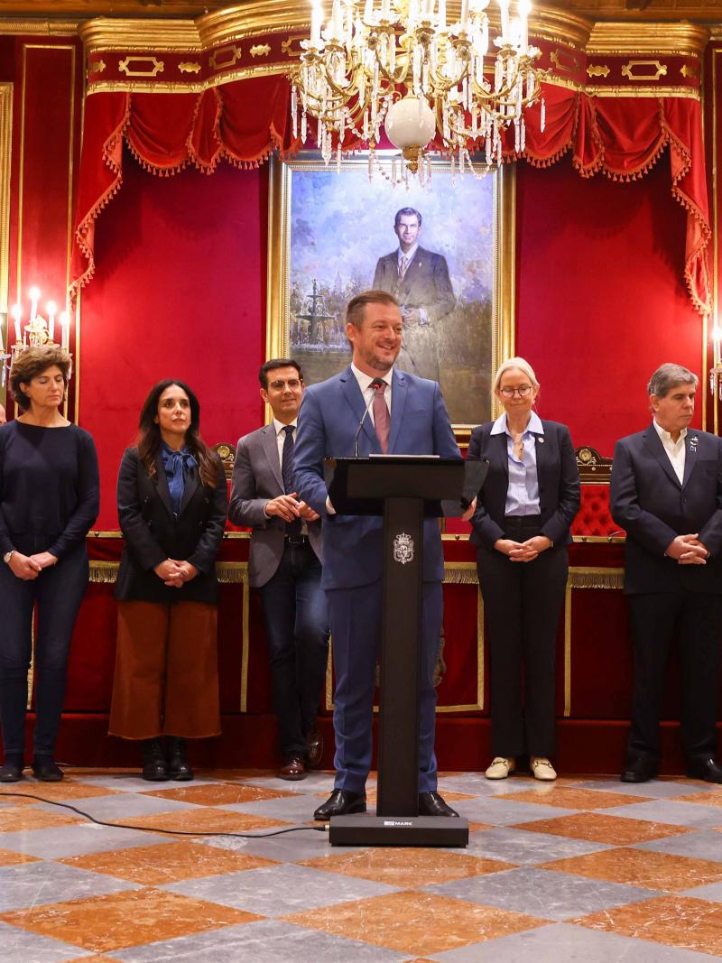 A man in a suit smiles while giving a speech at the podium in a lavishly decorated palace room.