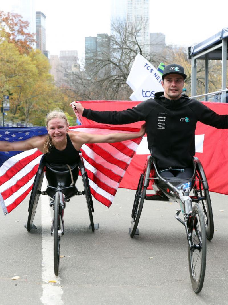 A female wheelchair racer with the USA flag and a male racer with the Swiss flag in front of a marathon finishing line