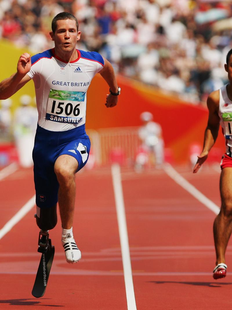 Two men with prosthetic legs sprint on an athletics track at a packed stadium.