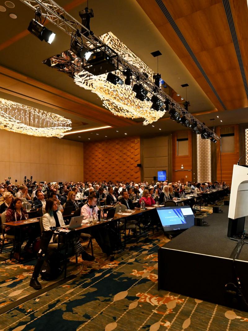 A man gives a speech from the stage as a room full of IPC member organisations looks on.