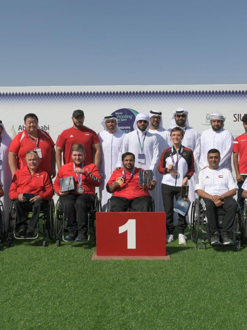 A group of nine men in wheelchairs posing in a podium with 14 people behind them