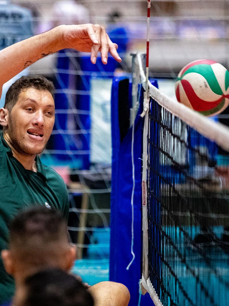 A male sitting volleyball player raises his right arm to hit the ball above the net