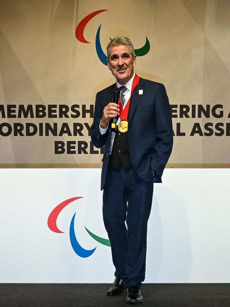 A man with a gold medal around his neck gives a speech from the stage with the Agitos logo behind him.