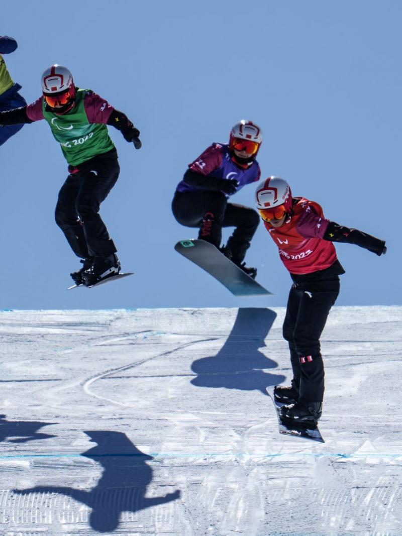 Four male snowboarders, all wearing vests that say Beijing 2022, helmets and goggles, compete at the Beijing 2022 Paralympic Games