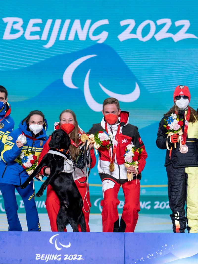 Six athletes pose for a photograph on the podium. A female athlete holds a gold medal with her left hand and her black guide dog with her right hand.