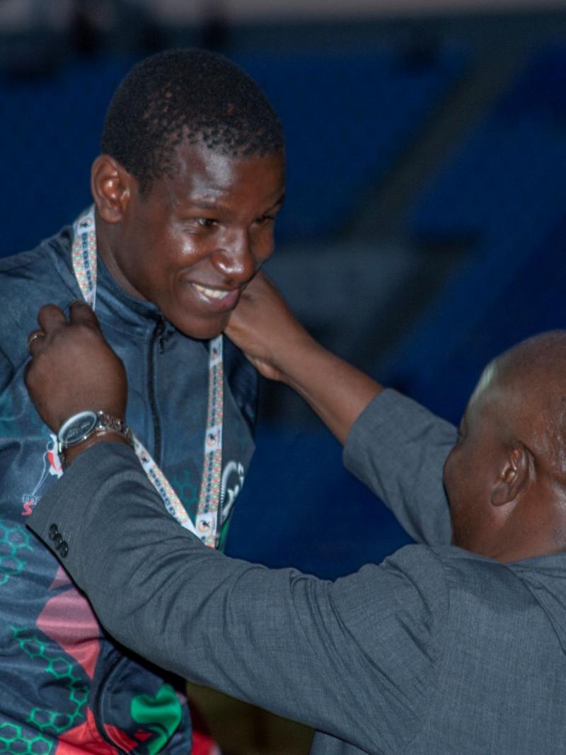 A male athlete in a national kit smiles as a medal presenter puts a medal around his neck.