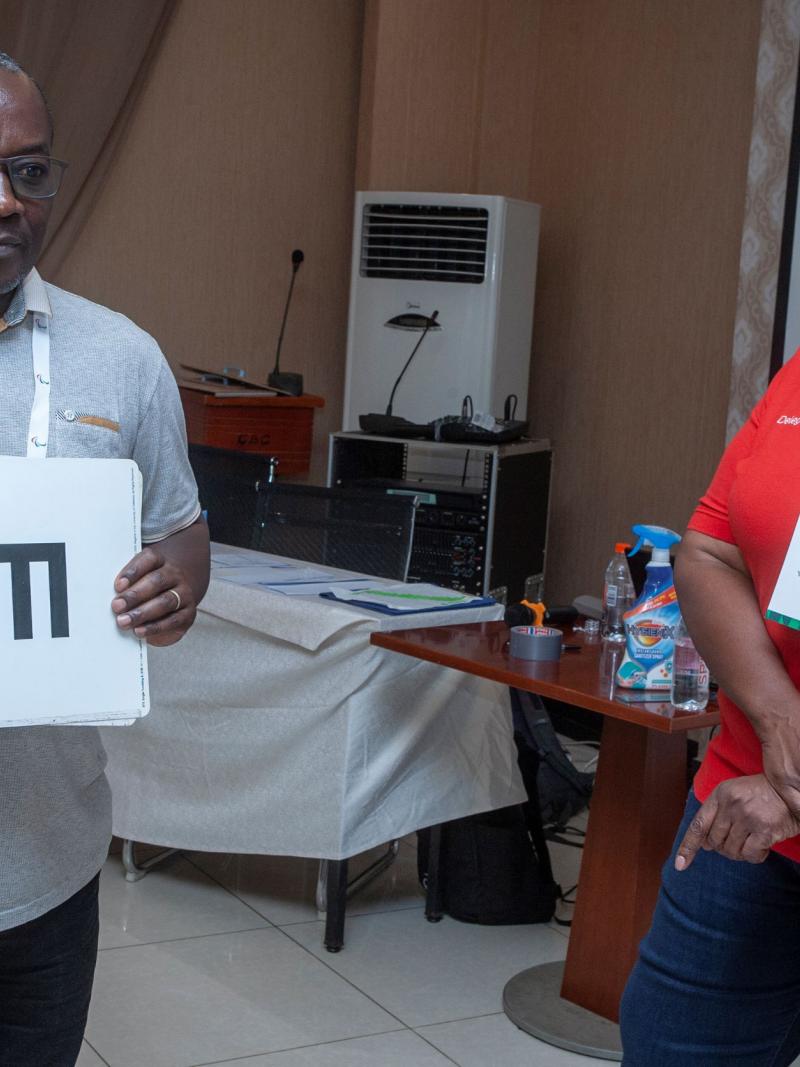 A man holds up charts for testing eyesight while a female supervisor looks on.