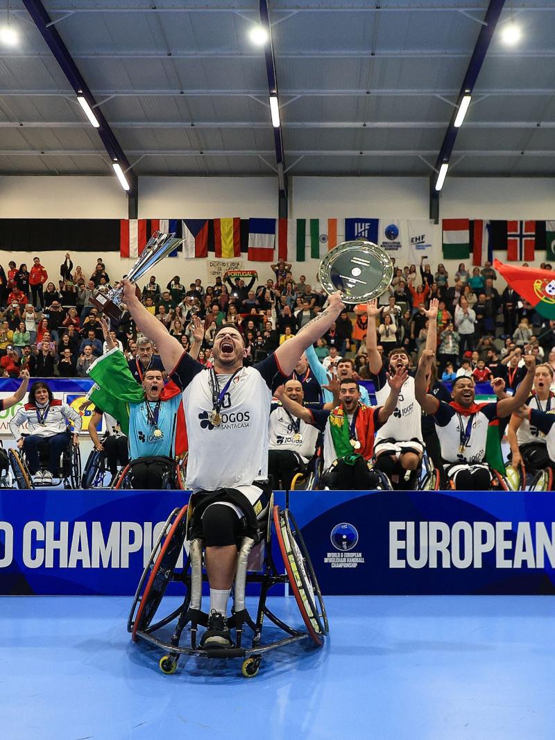 A male athlete in a wheelchair screams in celebration while holding up a trophy and dish, as a team cheers behind him next to the banner "world champions" and "European champions".