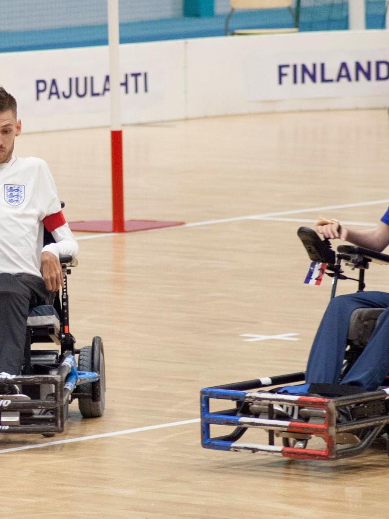 A male powerchair football athlete controls his wheelchair in front of the ball, while another player looks at the ball