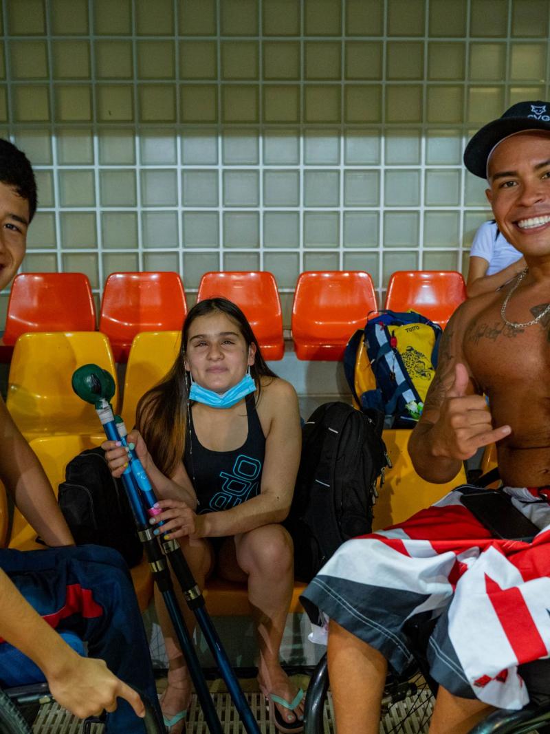 Three swimmers, two males in wheelchairs and a female swimmer holding equipment for athletes with vision impairments, pose for a photo on the bleachers.