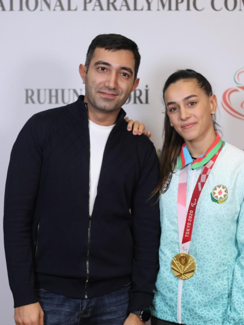 A man in and a woman, her right hand on his shoulder and a gold medal around her neck, pose in front of an official backdrop.