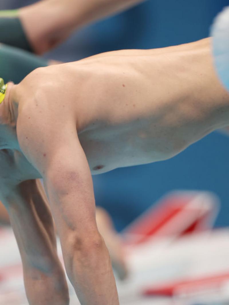 A male swimmer with a cap of Australia jumping in the water with another male swimmer in the foreground