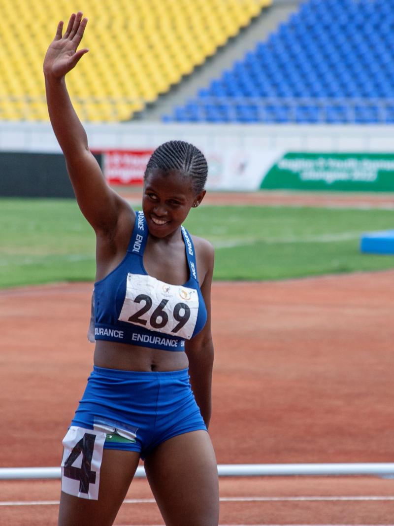 A female runner smiles and raises her right arm in a salute as she comes off the running track.