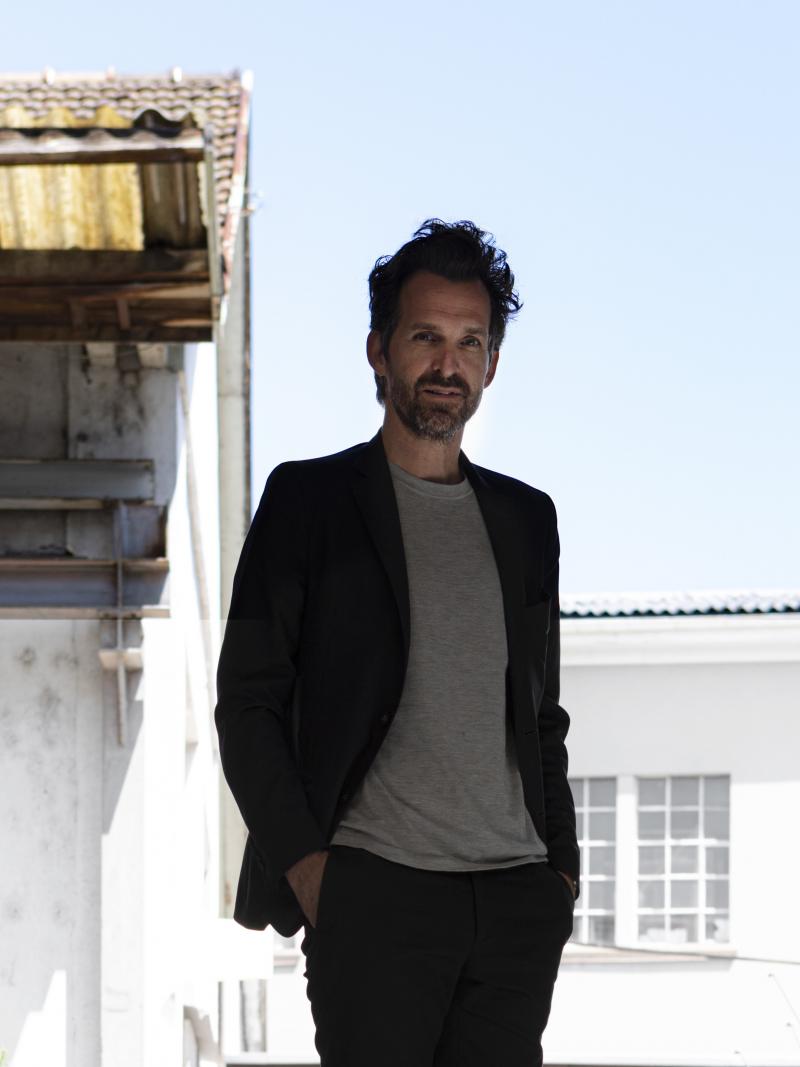 A man in a T-shirt and black suit poses with the backdrop of a slightly dilapidated house.