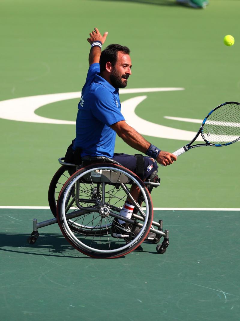 A male wheelchair tennis player makes a serve on a hard green court with his left arm stretched out wide.