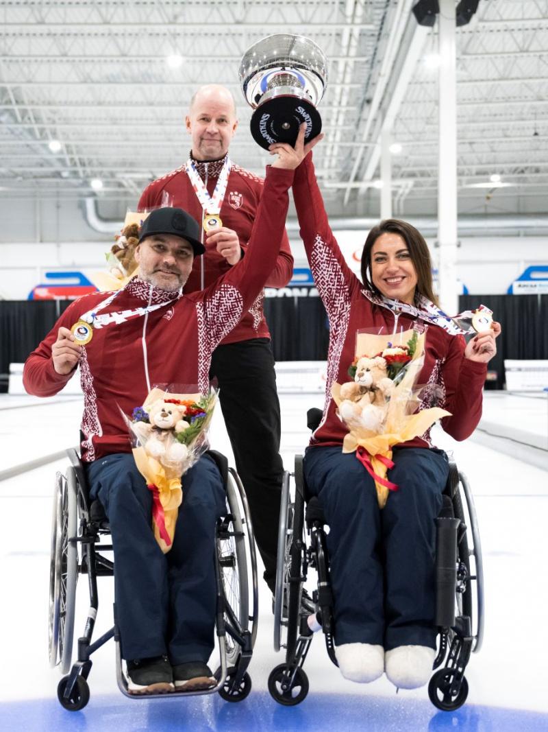 A female wheelchair curler and a male wheelchair curler lift a trophy together, while showing a gold medal
