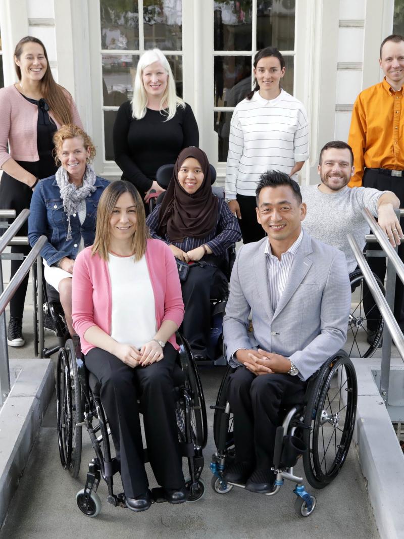 Nine athletes, five in wheelchairs at the front and second rows, and four standing in the back pose for a photo in front of the IPC headquarters.