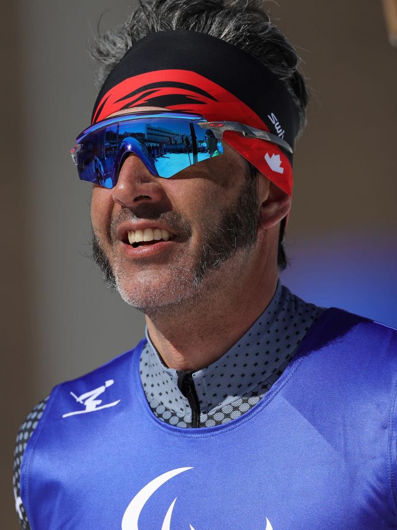 A close-up of a male cross-country skier in sunglasses and competition bib, holding skis in one hand. 