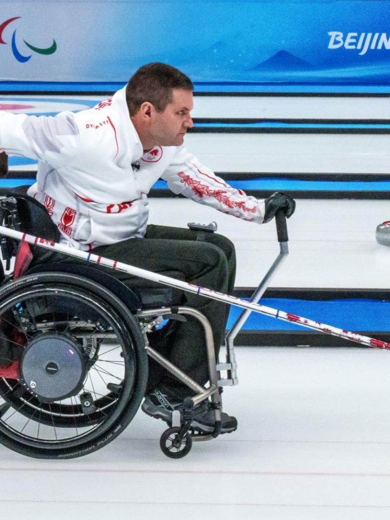 A male athlete slides a curling stone using a stick on the ice at Beijing 2022. He is on a wheelchair and another male athlete is holding onto the tires.