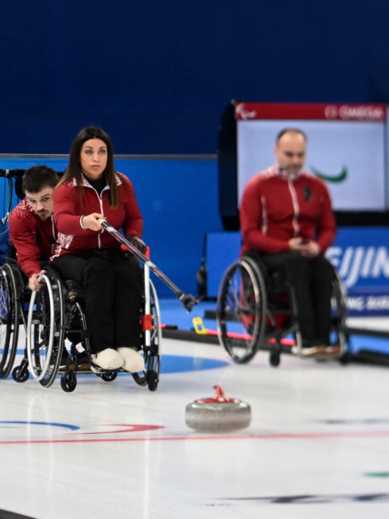 A female wheelchair curler delivers a stone with a stick, while a male athlete holds the wheels of her wheelchair.